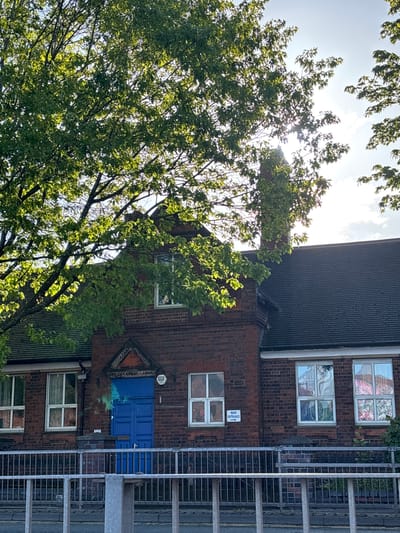 A daytime view of a building, likely a school in Newcastle-under-Lyme, United Kingdom, partially obscured by a tree. The building is constructed of red brick with a dark, corrugated roof. There are multiple windows, some with patterned coverings or artwork inside. One window displays what appears to be stained glass. There is a blue door with a triangular architectural feature above it. A fire escape ladder is visible on the right side of the building. A sign, partially obscured, is visible near one of the windows. The sun is shining through the tree branches.