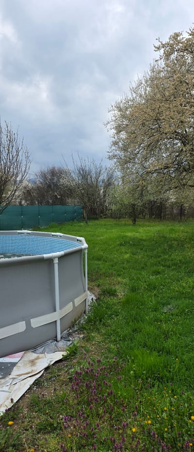 The image depicts an above-ground swimming pool situated in a yard. The pool is gray with a blue tiled interior pattern visible. A white frame supports the pool. The yard is grassy, with areas of green lawn and patches of mixed grass with purple and yellow wildflowers. Trees line the background. A green mesh fence is visible behind the pool. The sky is overcast with gray clouds. The location is Novachene, Bulgaria.