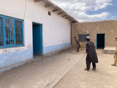 The image shows three boys playing cricket in a courtyard in Saranan, Pakistan (latitude: 30.532, longitude: 66.909). The courtyard is surrounded by whitewashed buildings with blue trim. There is a blue door on the left side of the image and a window above it. On the right side of the image is a building with a blue door and window. There is a red cricket ball in the center of the image. One of the boys is wearing a black long-sleeve shirt and black pants. He is holding a bat and is facing the camera. Another boy is wearing a brown shirt and pants. He is holding a yellow bucket and is facing the opposite direction of the first boy. The third boy is wearing a long white shirt and brown pants. He is facing the camera. There is a blue sky with white clouds in the background.