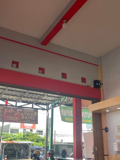 The media file depicts a street scene in Tampingan, Indonesia, as indicated by the location context. The image is shot from a slightly low angle, looking upwards towards a building's facade. The primary subjects are architectural elements and signage.

The building features a predominantly white and grey exterior with red accents, including a prominent red beam running diagonally across the frame. There are several square openings on the wall, some with recessed metallic squares inside. A white light fixture is visible on the left side of the frame, attached to the ceiling. Further up, a black device, possibly a security camera or projector, is mounted on a wooden beam.

In the background, a metal structure with horizontal slats suggests a storefront or covered area. Several motorcycles and their helmets are parked, hinting at a busy street or commercial area. Green trees are visible in the mid-ground, adding a touch of nature to the urban environment.

Several signs are present, though some are partially obscured or upside down. One large banner, oriented vertically, displays text in a bold, yellow font with a crown graphic. Text visible on this banner includes "AT2SAM3" and what appears to be "PESTAA HARMONI" and "CHICKEN ROCKET". Another sign in orange has the text "KARYAGUNA". The lighting suggests it is daytime, with sunlight filtering through, creating some shadows. The overall impression is of a vibrant commercial district.