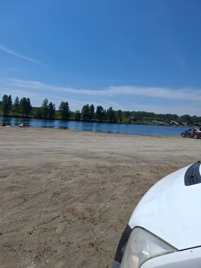 The image depicts a sunny day at a body of water, likely a river or lake, in Verkh-Katunskoye, Russia. The foreground is a dirt or gravel area with tire tracks and sparse vegetation. A white vehicle's hood and headlight are visible in the lower right corner. In the middle ground, several people are sunbathing or relaxing on towels near the water's edge. The background features a dense line of trees along the opposite bank of the water, with some structures, possibly houses or cabins, visible amongst the trees. Further in the background, more trees and a bright blue sky with wispy clouds complete the scene. Several cars are parked near the water.