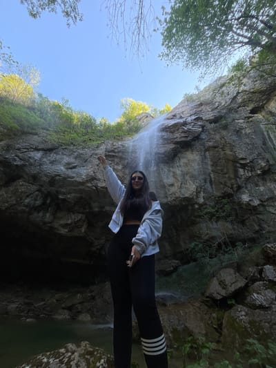 A person stands near a waterfall in a rocky environment. The location is Veselinovo, Bulgaria.
The person, presumably a woman, is wearing sunglasses, a light-colored cropped jacket, a dark-colored top, black leggings, and what appears to be leggings with stripes near the lower legs. She is pointing with her right hand towards the rocky wall beside the waterfall.
The waterfall cascades down a tall, craggy rock face, partially covered with greenery. Above the rock face is a blue sky with some trees.
The composition suggests a natural setting with emphasis on geological formations and vegetation, typical of Veselinovo, Bulgaria.