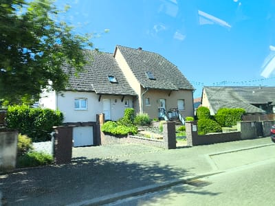 A two-story house with a gray tiled roof stands next to a similar structure. The first house has a white garage door and a small window above it. A white door with a small window is next to the garage. The second house features a set of stairs leading to the front door. Low brick walls define the property lines, adorned with greenery. A street runs in front of the houses.