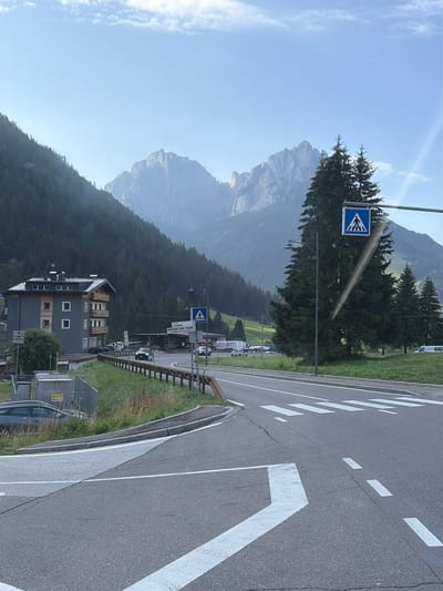 The image shows a road scene in the vicinity of Pera, Italy. In the foreground, a paved road with white lane markings and a crosswalk is visible, with a slight incline. To the left, a grey car is parked alongside a grassy embankment and a fence enclosing some electrical equipment. A modern, multi-story building with a grey facade and wooden accents is situated on a hillside to the left of the road. Behind the building, a dense forest covers a steep mountain slope.
In the middle ground, a two-lane road curves to the right. Several cars are present on the road, including a dark colored vehicle driving towards the viewer and a white van parked in the distance. A pedestrian crossing sign with a blue background and a white pictogram of a person walking is mounted on a pole, to the right of the road. Further to the right, a cluster of tall evergreen trees stands against a backdrop of rugged, mist-shrouded mountains. A street lamp is also visible near the pedestrian crossing sign. The sky above is a pale blue with scattered clouds. A signpost on the right side of the road indicates "Strada JUMETA ADVENTURE PARK".
