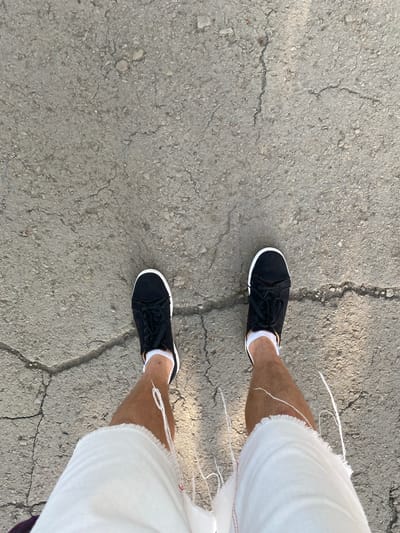 The image is an overhead view of a person's legs and feet standing on a cracked asphalt surface. The person is wearing black sneakers with white soles and white socks, and baggy white shorts with frayed edges. The asphalt has several visible cracks and is textured with small stones and debris. The lighting suggests it is daytime, with some shadows cast on the ground. The location is identified as Antratsyt, Ukraine.