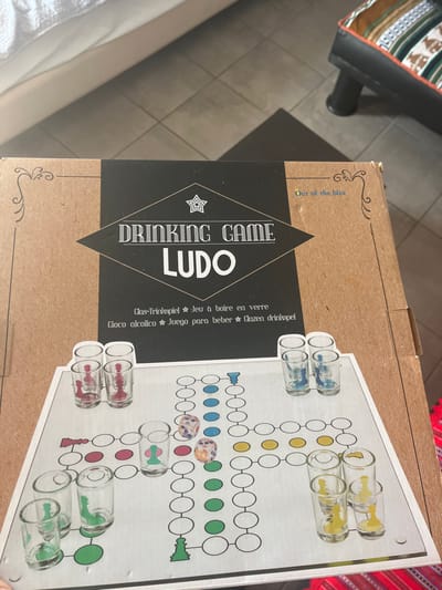 The image shows a cardboard box on a tile floor, likely indoors, with the lid slightly open. The box contains a "Drinking Game Ludo" game, designed for playing with shot glasses. The box is made of brown cardboard and has a black and white label with the game name and a depiction of a game board featuring a simplified version of Ludo, with a black and white checkered pattern, coloured circles, and five shot glasses arranged in a row on the top left side of the board, with a star in the center. The box is lying on a white bed with a white blanket. There is an orange and black patterned rug on the floor to the right. This is a common game, found in many locations, but the coordinates provided, 40.627, 24.575, locate the image in Limenaria, a small town on the island of Thasos, Greece. The box is the only object present in the image. There are no people in the image.