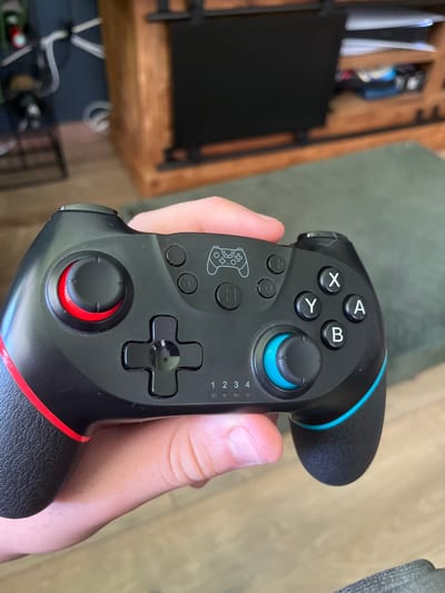 A close-up, eye-level shot shows a person's hand holding a black and red video game controller. The controller features a directional pad, four face buttons labeled X, Y, B, and A, two analog sticks, and several smaller buttons. The right analog stick has a bright blue accent, and the controller has thin blue and red trim. The background is softly blurred, with hints of wooden furniture and a textured green surface. The lighting appears to be natural, suggesting daytime. The image is focused on the controller, highlighting its design and buttons.