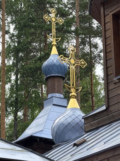 The image shows the exterior of a wooden church with blue, diamond-patterned domes, topped with golden Orthodox crosses. The church is surrounded by tall, green pine trees, and the sky is overcast. The wooden structure of the church is visible on the right side of the frame, with horizontal planks and a dark wooden trim. The roofing is made of gray metal panels. The scene is located in the village of Nenyalki, Leningrad Oblast, Russia. There are no people visible in the image, and no actions or interactions are taking place.