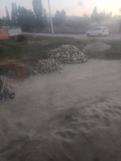 The image depicts an outdoor scene, likely a construction or work area, in Кара-Буура, Kyrgyzstan. In the foreground, there is a large expanse of sandy ground with tire tracks visible, suggesting recent vehicle activity. To the left of the sandy area, a pile of loose straw or dry grass is visible, adjacent to a pile of stones. Further to the left, leaning against a surface, are two shovel-like tools. In the mid-ground, there are two piles of stones, one larger and more circular than the other. Behind the stones, a light-colored van is parked on a road. To the far left, an orange and white truck with a flatbed is parked. The background shows a line of tall, slender trees and some hazy structures, possibly buildings. The sky is overcast and appears to be either early morning or late evening, with soft, diffused light. The overall impression is one of a utilitarian or undeveloped area.