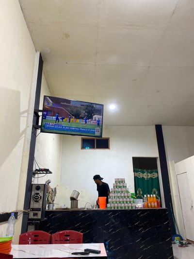 The image depicts an indoor scene, possibly a small eatery or shop, in Luengbaro, Indonesia. A man wearing a black t-shirt and a black cap is standing behind a counter. On the counter, there are stacks of canned goods, bottles of liquid, a jug, and some eggs. To the left of the man, a large speaker system is mounted on a wall. Above the counter, a television screen displays a soccer game with Indonesian subtitles. The walls are painted white, and the ceiling appears to be made of concrete with some water stains. In the foreground, red chairs and a table are visible, suggesting a seating area. The overall environment appears functional rather than decorative.