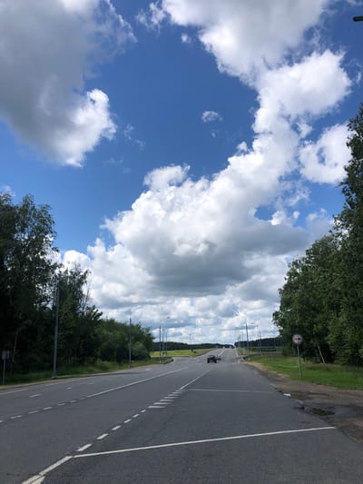 A view of a paved two-lane highway under a bright blue sky with scattered white cumulus clouds. The road curves to the right in the distance. Trees line both sides of the road, with the foliage appearing dense and green. Several streetlights are visible along the roadside. In the distance, a dark-colored car is traveling on the highway. A traffic sign showing two cars side-by-side within a red circle is posted on the right side of the road. There is a pothole filled with water on the shoulder of the road. The scene appears to be in a rural or suburban area. The location is identified as Vysotnik, Russia.