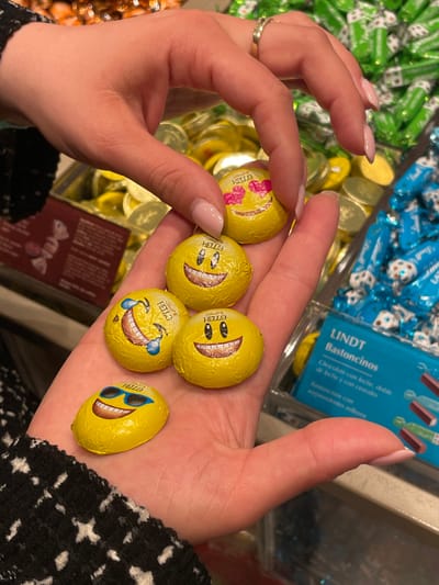 A close-up photograph captures two hands interacting with five emoji-themed chocolate candies within a retail environment in Marratxí, Spain. The upper hand, adorned with a gold ring on the ring finger and light-colored nail polish, delicately holds one circular yellow-foiled chocolate depicting a "heart eyes" emoji and the text "HELLO." The lower hand, open with its palm up and also displaying light-colored nail polish, cradles four additional yellow-foiled chocolates. These include one circular "grinning face with smiling eyes" emoji, one circular "face with tears of joy" emoji, another circular "grinning face with smiling eyes" emoji, and an oval-shaped "smiling face with sunglasses" emoji, all bearing the "HELLO" text. In the blurred background, various bins or displays of wrapped candies are visible, including green-wrapped items to the upper right, gold-foiled circular chocolates, and a transparent container on the right holding blue-wrapped Lindt Bastoncitos. A dark red sign with white text is partially visible on the left side of the frame.