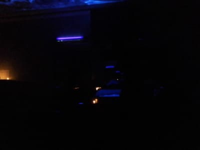 The image depicts an extremely dark, low-light environment with limited discernible detail. Sparse artificial light sources provide the primary illumination. A prominent, elongated horizontal band of vivid purple-blue light is centrally located in the upper portion of the frame. Below this, multiple smaller, less defined points of blue light are scattered across the lower and central areas. In the extreme lower-left quadrant, a single, faint, warm orange light source is visible, casting minimal localized illumination. No identifiable individuals, objects, or specific architectural features are visible due to the severe underexposure. The precise nature of the environment (e.g., interior, exterior, specific establishment) cannot be determined. The image content does not contain sufficient visual information to identify a specific location within the city of Long Beach, United States. No actions or interactions are observable.