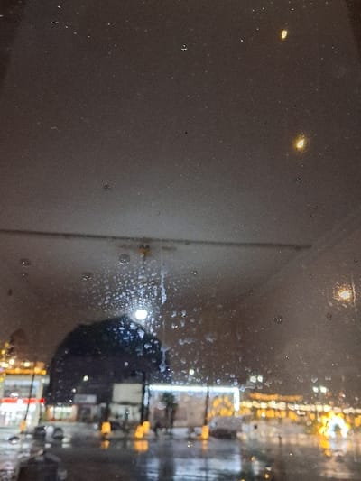 The image displays an exterior nighttime urban scene in Lanchkhuti (ქალაქი ლანჩხუთი), Georgia, captured through a wet and partially obscured transparent barrier, likely a window. The immediate foreground is characterized by numerous irregularly shaped water droplets, smudges, and fine particulate matter adhering to the viewing surface. A faint, horizontal, light-colored linear reflection, possibly originating from an interior overhead light fixture, is visible across the upper-middle section.
The background reveals a wet city street or public square, suggesting recent or ongoing rainfall, evidenced by the highly reflective ground surface. Multiple sources of artificial light illuminate the scene: diffuse yellow-orange streetlights, brighter white and red lights from commercial establishments (including a prominent red and white illuminated element on the far left), and decorative yellow/gold lights, some forming what appears to be an archway or string lights on the right side. These lights generate distinct reflections on the saturated ground. Indistinct building structures are visible, with one exhibiting a rectangular white light outline. A large, dark, amorphous shape is prominent in the central-left area, partially obscuring the background. No specific individuals or clearly defined vehicles are discernible. The overall environment is one of an illuminated urban setting during inclement weather at night.