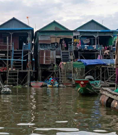 This image captures a stilt village on the water in the city of Soutr Nikom, Cambodia. The village consists of several houses built on wooden stilts, with many appearing weathered and constructed from corrugated metal and wood. The houses are multi-story, with open-air walkways and balconies. Laundry is visible hanging from some of the structures.

In the foreground, a body of water reflects the sky and the stilt houses. Several small boats are present, some tied to the houses and others in the water. One boat in the middle ground appears to have people in it, including someone steering with an outboard motor. Another boat, painted green and red, is moored to a floating platform made of barrels.

There are people visible on the walkways and stairs of the houses, suggesting ongoing daily activity. Some individuals are standing, while others are seated. The overall scene depicts a densely populated community living directly on the water, with a sense of both vibrancy and the harsh realities of life in such an environment. The sky is overcast, contributing to a diffused light across the scene.