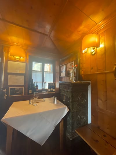 The image depicts a cozy dining area, likely within a restaurant in Alagna Valsesia, Italy. A table draped with a white tablecloth is set for two, with wine glasses, cutlery, and salt and pepper shakers. To the right of the table, a decorative tiled stove stands against a wood-paneled wall. Above the stove, a vase holds a few red flowers. A wooden bench runs along the wall to the right. On the left wall, two framed certificates and a framed photograph are displayed. Through a window, partially obscured by white curtains, glimpses of buildings can be seen. The warm lighting, likely from wall-mounted lamps, casts a golden hue over the wooden interior, creating an intimate atmosphere.