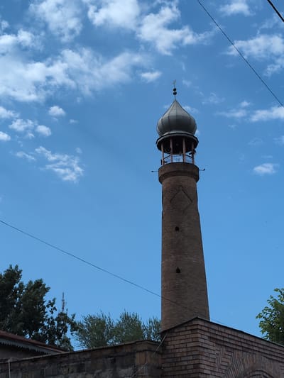 The media file depicts a minaret against a bright blue sky with scattered white clouds. The minaret is constructed from dark bricks and has a conical, metallic dome at its apex. The lower portion of the minaret features a circular opening with intricate latticework or decorative panels. A thin wire or cable runs diagonally across the frame, partially obscuring the sky and the minaret. To the right of the minaret, the corner of a brick building is visible, also appearing to be constructed from similar dark bricks and featuring arched architectural elements. Green foliage from trees is present at the top and bottom right of the frame, suggesting an outdoor setting. The perspective is from a low angle, looking upwards towards the minaret and the sky. The lighting indicates it is daytime, likely late afternoon or early evening given the quality of the light and the slight warmth in the sky. There are no people visible in the frame, and no discernable text. The scene evokes a sense of historical architecture and a clear, pleasant day. The minaret's design suggests it is likely part of a mosque or a similar religious or historical structure in Şəki, Azerbaijan.