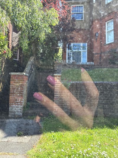 The image shows a view through a window, reflecting a hand with painted nails. The backdrop visible through the window is a residential scene in Guildford, United Kingdom. The buildings are predominantly brick, with visible windows and a white door. Lush greenery, including trees and a grass verge with daisies, is present. A brick wall is also visible in the yard, adjacent to a sidewalk. The scene appears to be taken on a sunny day.