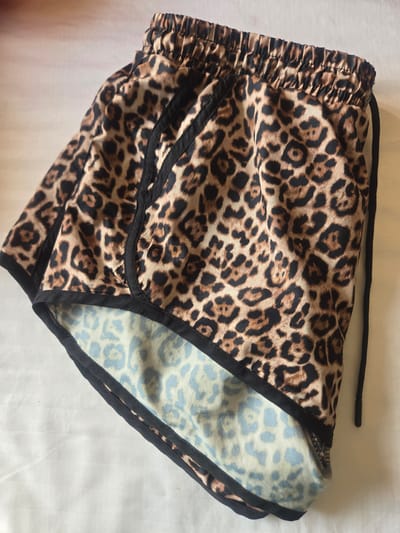 The image displays a pair of leopard-print athletic shorts laid flat on a white, lightly textured surface. The shorts are a light brown color with black spots, and they have black trim along the edges and the waistband. The waistband appears to be elasticized, and a black drawstring is visible hanging from the side. The inside lining of the shorts is visible at the bottom, revealing a lighter leopard print in shades of blue and white. The lighting suggests an indoor setting. There is no indication of a city location or the United Arab Emirates.