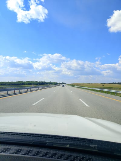 The image depicts a view from the driver's seat of a vehicle traveling on a highway. The highway is paved with asphalt and has clear white lane markings. A yellow line separates the lanes of traffic. On the left side of the highway, there is a metal guardrail with orange and white reflective markers. Beyond the guardrail, there is a green treeline that stretches into the distance. The landscape on the right side of the highway is a rolling field of dry grass, with scattered trees visible on the horizon. The sky is a bright blue with scattered white cumulus clouds. Several vehicles are visible on the highway in the distance, moving in the same direction as the camera's vehicle. The foreground shows the white hood of the vehicle and a black dashboard. The image appears to have been taken during the daytime under clear weather conditions. The location is identified as the highway within Алтынды ауылдық округі, Kazakhstan.