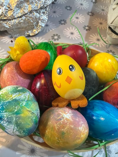 The image shows a collection of Easter eggs in a basket. The eggs are dyed in various colors, including yellow, red, blue, orange, green, and iridescent combinations. There is a yellow egg with a face drawn on it and brown felt ears attached. Green paper grass is used as filler around the eggs. The background is a tablecloth with a floral pattern. To the left, foil wrapping is visible. There is no discernible information to determine the location as Enniskillen, United Kingdom. The objects are still life. No people, actions, or interactions are visible.