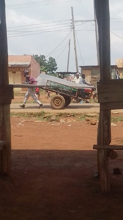 The scene is set in Siaya Township ward, Kenya, viewed from a slightly elevated or framed position overlooking a paved road and its dirt shoulders. In the midground, a red motorbike pulls a two-wheeled cart laden with cargo, moving from left to right. The cart carries two large, reflective, rectangular sheets, possibly corrugated iron, extending significantly beyond the cart's frame, along with green netting and a green jerry can. Two males occupy the motorbike and cart: one dark-skinned male in a grey hooded sweatshirt with "97" visible, driving the motorbike, and an older, light-skinned male in a light shirt and cap, seated on the cart behind him, smiling.
Parallel to the motorbike and cart, on the left, a light-skinned male wearing light grey trousers, dark shoes, a dark cap, and a red-patterned cloth is walking, also moving left to right. He carries a long, reflective, metallic object horizontally in front of him.
The immediate foreground consists of reddish-brown dirt, with dark wooden posts and planks framing the view on the left and right. In the background, to the left, is a light brown building with a sign indicating "DETERGENTS" and "GRACE SHOP." To the right, various structures with corrugated metal roofs and walls are visible. Overhead, multiple wooden utility poles with numerous power lines crisscross against a light, overcast sky.