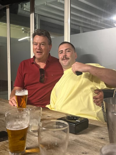 Two adult males are depicted seated at a wooden table within an establishment in Bellambi, Australia, during evening hours. The male on the left, appearing older, has short, dark hair and wears a dark red polo shirt with sunglasses hanging from the collar. He is looking towards the camera with his mouth open. His right hand rests near a glass containing an amber liquid consistent with beer, while his left arm is positioned around the shoulder of the male on the right. The male on the right, appearing younger, has short buzzed dark hair and a mustache. He wears a loose light yellow t-shirt and looks towards the camera with a slight smile, his teeth visible. He holds a dark rectangular object in his raised right hand, near his shoulder, and his left hand is placed on the older male's back. On the rustic wooden table, two glasses contain amber liquid, while two other clear glasses appear empty. A black rectangular object, possibly an ashtray, and a partially visible dark smartphone are also present on the table. A silver metal container is partially visible on the far right. The background consists of large glass panels with horizontal frosted sections, behind which a white security camera is mounted in the upper left. The scene is illuminated by artificial lighting.