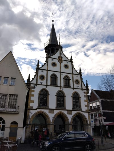 An external view of the historic Rathaus (Town Hall) in Burgsteinfurt, Germany, is depicted under a partly cloudy sky. The multi-story central building features a white facade with dark stone and trim accents, characterized by a prominent stepped gable with decorative finials. A steep, dark conical spire, topped with a weathervane (featuring a cardinal direction indicator and an animal figure), rises from the roofline. The facade displays a golden coat of arms or emblem on its upper section, along with several arched windows on the lower levels and circular windows on the upper levels, all with dark frames. Bird-deterrent spikes are visible along ledges. The ground floor exhibits multiple large stone arches.
To the left, an adjacent light-colored stucco building with a simpler gable roof features multiple rectangular windows and a second-story balcony with an ornate dark metal railing. Outdoor cafe-style tables and chairs are positioned in the foreground near this building.
To the right, a darker building with a visible reddish-brown roof and white-framed windows is partially observed. A dark street pole with multiple white and red directional signs, including one for "Fußgängerzone" (pedestrian zone) and town names such as "Horstmar," stands near this building. A red umbrella from an outdoor seating area is also partially visible.
At street level, a dark-colored SUV is parked parallel to the curb in front of the Rathaus. Several individuals are present on the paved surface near the Rathaus entrance. This includes an adult dressed in dark clothing walking alongside a child in a pink jacket, and another adult carrying a red backpack. The ground surface appears to be paved. Bare tree branches are visible on the far right, suggesting a late autumn or winter season.