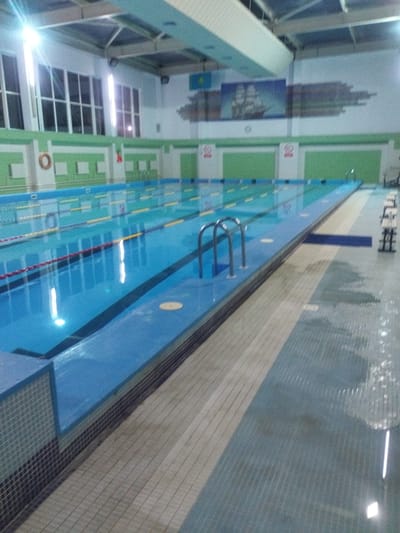 The image depicts an indoor swimming pool in Қотырбұлақ, Kazakhstan. The pool features multiple lanes marked with lane ropes and yellow buoys. The water is a clear blue. The pool area is surrounded by blue-tiled edges and a beige tiled walkway. Along the wall, there are green and white decorative elements, including a life preserver ring and signs with a red circle. A mural of a sailing ship is visible on the wall above the pool. Natural light enters through large windows, and artificial lights illuminate the space. The floor surrounding the pool appears wet in some areas. There are no people visible in the image.