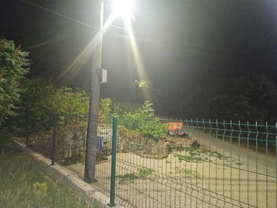 The image depicts a nighttime scene on a road in Zlataritsa, Bulgaria. A bright street lamp illuminates the area, casting long shadows and creating lens flares. In the foreground, a green wire mesh fence runs horizontally across the frame, bordered by a concrete curb and tall grass. To the left, a concrete utility pole with attached electrical boxes and wires stands prominently. Beyond the fence, the terrain slopes upwards, with dense vegetation and scattered rocks. A small orange utility vehicle with its brake lights illuminated is visible on the road in the background, appearing to be parked or moving slowly uphill. The overall environment suggests an outdoor, possibly rural or semi-rural, setting at night.