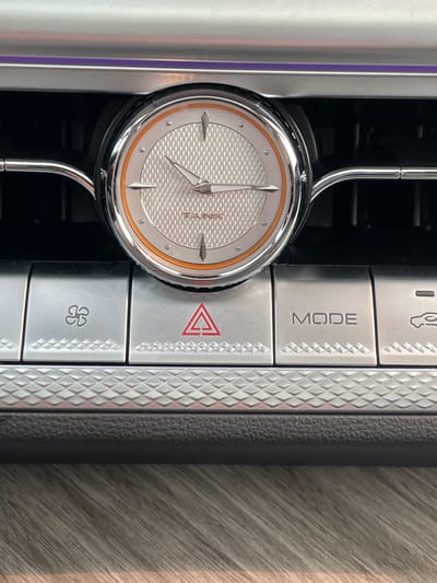 A close-up, head-on shot shows the interior of a vehicle, focusing on a circular analog clock with a white textured face and orange accents, centrally positioned above a row of silver buttons. The clock is framed by a chrome bezel and is integrated into a dark grille. Below the clock, three buttons are visible: the leftmost button displays a fan icon, the middle button features a red hazard triangle symbol, and the rightmost button is labeled "MODE." The buttons have a textured diamond pattern along their lower edges. The overall setting suggests a modern car interior, with a hint of wood-grain texture visible at the bottom of the frame. The location is not specified in the image.
