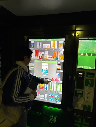 The image shows an outdoor pharmacy vending machine at night. The machine displays various pharmaceutical products, with their prices and product codes. A person, partially visible, is interacting with the touchscreen interface of the vending machine, their finger pointed towards a selection. The vending machine has a prominent "PharmaShop24" logo. To the right of the main vending machine, another interactive screen is visible, displaying options like "Compra" (buy), "Chiamata" (call), and "Ritiro" (pickup). Payment options including a card reader and coin slot are present. Instructions in Italian and English are posted on the machine. The background is dark, suggesting it's a nighttime scene in an urban or semi-urban environment. The location is identified as La Ila - Stern - La Villa, Italy.