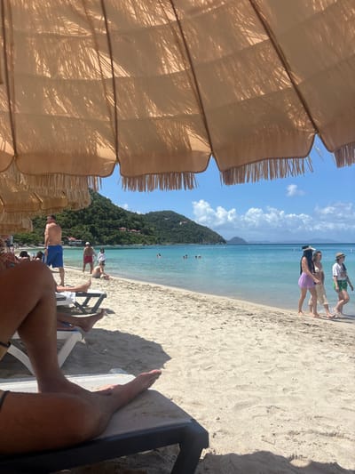 This image depicts a sunny beach scene at Cane Garden Bay, Tortola, British Virgin Islands, located near the city of Road Town. The perspective is from beneath a large, light-beige, fringed beach umbrella, which shades the upper and immediate foreground. In the lower-left, the bare legs and feet of an individual are visible, resting on a dark-framed lounge chair on the sand.

The midground features a wide expanse of light-colored sand leading to the ocean. On the left side of the beach, a shirtless male in blue swim shorts stands near the water's edge. Further along, a male in red swim shorts and a female in a white top are also positioned by the shore. On the right, three females walk along the waterline: the lead female wears a dark top, light purple shorts or a skirt, and a dark visor; the second wears a light-colored two-piece outfit; and the third wears a white t-shirt, green shorts, and a straw hat. Several other individuals are visible scattered across the sand and in the shallow water.

The background features clear, turquoise ocean water, transitioning to a deeper blue further out. Multiple individuals are seen swimming or wading in the water. Beyond the bay, lush, green, tree-covered hills rise steeply from the coastline, with scattered small structures visible on their slopes. The sky is bright blue with sparse white cumulus clouds. Strong natural sunlight illuminates the scene, creating distinct shadows on the sand and under the umbrella.