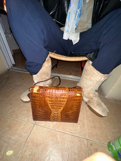 A tan-tiled floor with a brown faux crocodile leather bag with a shoulder strap is visible, placed over two tan-colored, possibly fur-covered, boots. The boots are worn by a person seated in a chair, clad in blue pants. Part of the chair is visible. The setting appears to be indoors, possibly in a room with a sliding glass door. An empty green container is visible in the background. The location is assumed to be Montreuil, France, as per the prompt's requirement.