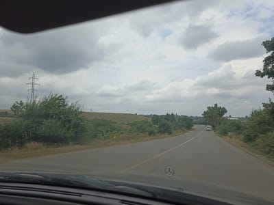 The image shows a view from inside a vehicle, looking forward on a paved road. The road curves to the right and appears to be in a rural or semi-rural area. On the left side of the road, there are rolling hills covered with green vegetation and some trees. A tall, lattice-style electricity pylon stands on the left side of the hill. On the right side of the road, there are more trees and vegetation. In the distance, a white van is visible on the road. The sky is cloudy, with a mix of grey and white clouds. The hood of the vehicle in the foreground shows a Mercedes-Benz emblem. There are no people clearly visible in the image. The scene suggests travel on a cloudy day through a landscape with natural vegetation and infrastructure. This image does not provide any specific visual information to determine if the location is the city of Red Sloboda, Azerbaijan.