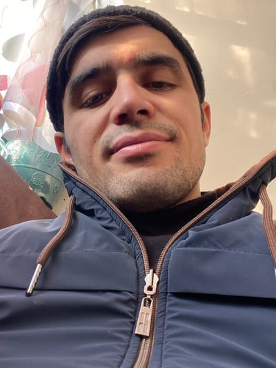 A man with light skin, wearing a black beanie, looks directly at the camera with a slight smile. His face has stubble, and his eyes are dark. He is wearing a blue zippered jacket with brown trim and metallic toggles on the drawstrings. The background is out of focus, but shows light and possible floral patterns. The image is oriented vertically and rotated slightly.