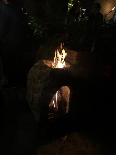 The image captures a nighttime scene in Santa Cruz Aquiahuac, Mexico, featuring a stone-built fire structure. This structure, composed of irregular light-colored stones and a reddish-brown brick section at its rear, contains two active fire points. An upper, circular opening emits prominent orange and yellow flames, functioning as a fire pit. A lower, arched opening reveals additional flames and burning logs or embers, suggesting an oven or fireplace. Dense green foliage is situated immediately behind the fire structure. The setting is dimly lit, with firelight providing the primary illumination. On the far left, the partial silhouette of an individual standing is visible, with dark shoes discernible. In the upper right background, two additional individuals are faintly present; one has dark hair and a possible green garment, while the other wears glasses and has short dark hair. No specific actions or interactions between the individuals are discernible due to the low light.