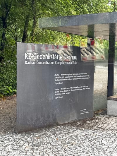This is a medium-shot, eye-level, outdoor photograph of the KZ-Gedenkstätte Dachau, the Dachau Concentration Camp Memorial Site, located in Dachau, Germany. The primary subject is a large, dark grey, rectangular monument with white text. The text at the top of the monument reads "KZ-Gedenkstätte Dachau" and below it, "Dachau Concentration Camp Memorial Site". Below this, there are two quotes, one in German and one in English. The German quote, attributed to Eugen Kogon, translates to: "Dachau – the significance of this name is from the German history. It stands for all concentration camps which the Nazis established in their territory." The English quote, also attributed to Eugen Kogon, is identical to the German translation.
Reflected in the glass canopy above the monument, there are images of what appear to be construction barriers or scaffolding. The background consists of lush green trees and foliage. The ground in front of the monument is paved with cobblestones. A small sign is visible in the lower right corner of the monument, which reads "KZ-Gedenkstätte Dachau Stiftung Bayerische Gedenkstätten". The overall lighting suggests an overcast day, and the monument appears slightly weathered. There are no people visible in the image.