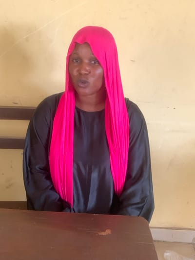 An adult female, approximately 30-40 years old, with dark skin, is depicted seated in an indoor setting. She is wearing a bright pink head covering that drapes over her shoulders and a long-sleeved black garment. Her mouth is slightly open, indicating an utterance or speech in progress. A dark wooden table occupies the lower portion of the frame, in front of the subject. The background consists of a plain, light-colored wall, likely tan or yellow, with a dark horizontal structural element visible behind her left shoulder. The scene is located in Abobo, Nigeria.