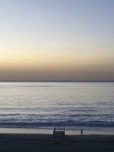 A serene coastal view. The sky transitions from pale blue to warm beige. The ocean stretches alongside, its surface textured with subtle wave patterns. A narrow beach borders the water. A lifeguard tower stands on the sand. A solitary figure walks along the shore, their silhouette visible against the water. The color palette is muted and cool, evoking a sense of calm.