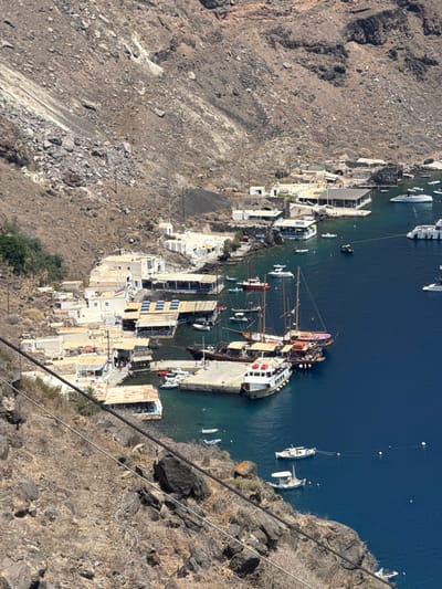 The image displays a high-angle view of the Old Port of Fira, also known as Skala, located within the Ia Municipal Unit on the island of Santorini, Greece. The scene is dominated by a deep blue bay nestled at the foot of a steep, arid, and rocky volcanic cliff face, which occupies the upper two-thirds of the frame. The cliff exhibits visible scree slopes, scattered dark volcanic rocks, and sparse, dry vegetation.
Along the narrow coastline at the base of the cliff, a cluster of whitewashed, flat-roofed buildings with traditional Cycladic architecture are visible. Many of these structures feature outdoor seating areas covered by awnings or pergolas, suggesting commercial establishments such as restaurants or shops. On the roofs of some buildings, dark rectangular panels, possibly solar installations, are present.
A prominent concrete pier extends into the bay, serving as a docking point. Moored at this pier are several large, multi-masted wooden sailing vessels, consistent with tourist excursion boats, and a substantial white passenger ferry or tour boat. Numerous smaller vessels, including white motorboats and dinghies, are scattered throughout the tranquil blue waters of the bay, some anchored or moored closer to the shoreline. One small boat appears to be underway near the pier.
Two thick black cables, indicative of a cable car system, run diagonally across the lower left foreground, descending towards the port area from an unseen elevated position. Small, indistinct human figures are discernible on the pier and near the waterfront buildings, suggesting general human presence and activity within the port. The overall lighting indicates a clear, sunny day.