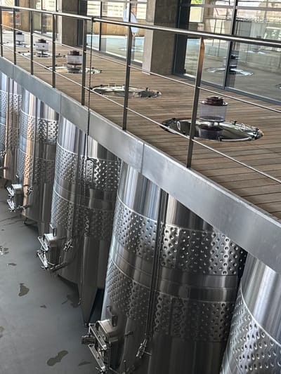 The image shows the interior of a winery in Mukhrani, Georgia. In the foreground, several large, stainless steel fermentation tanks are lined up. The tanks have a textured surface with many small, raised circles. Each tank has a complex valve system at its base. Above the tanks, a wooden deck is visible, with a metal railing running along its edge. Several circular lids with glass or plastic components are set into the deck. The background features large windows and concrete pillars, suggesting a modern industrial setting. There are no people visible in the image.