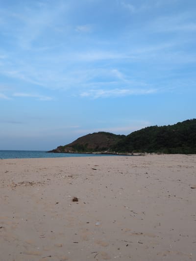 The image depicts a serene beach scene, likely within Hoành Sơn Ward, Vietnam. A wide expanse of light brown sand stretches across the foreground, scattered with small shells, twigs, and other debris. Gentle waves of turquoise water meet the shore, leading the eye to two tree-covered hills in the distance. The larger, left hill is dominated by sparse brown vegetation, while the right hill is densely covered in dark green trees. The sky above is a pale blue, streaked with wispy white clouds. There is a solitary, faint human figure visible in the water on the far left of the image. The overall impression is one of a peaceful, natural coastal environment.