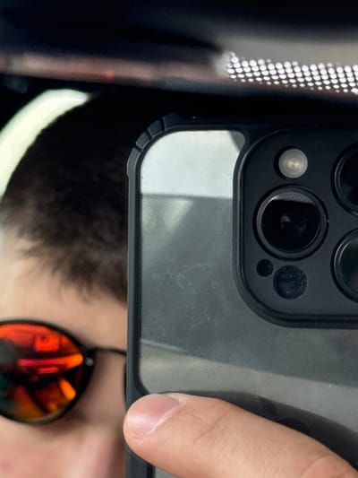 The scene depicts a close-up, reflected image of an individual holding a smartphone within what appears to be a vehicle interior. The location is Gorski Izvor, Bulgaria.

**People:** One individual is partially visible in reflection. The upper left quadrant shows the crown and back of the head, with short, dark hair. The face is largely obscured by dark-framed sunglasses with reflective red-orange tinted lenses. No distinct facial features are identifiable. A human hand, likely the individual's right, is visible on the right side of the frame, with the thumb pressed against the side of a smartphone.

**Subjects/Objects:** The primary subject is a dark-colored smartphone, appearing to be an iPhone model, encased in a textured, dark protective cover. The phone's rear camera array, consisting of three prominent lenses, a flash unit, and a smaller sensor, is clearly visible. The surface of the phone case shows minor smudges and dust. The reflection also includes a car's overhead light fixture, characterized by a horizontal strip of small, uniformly arranged circular LED lights.

**Actions/Interactions:** The individual is actively holding the smartphone to capture a reflection, indicating the deliberate act of photographing the mirrored image.

**Environment:** The setting is indoors, specifically within the confined space of a vehicle, evidenced by the characteristic overhead LED lighting and the general composition suggesting a car's interior or rearview mirror reflection.