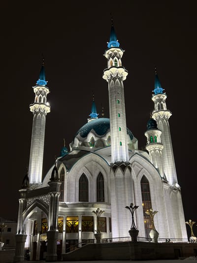 The image depicts the Kul Sharif Mosque, brightly illuminated at night, located in Tashevka, Russia. The grand structure features a central main dome of a blue-green hue, flanked by four tall, slender minarets with matching blue-green conical spires, two of which are prominently visible. Two smaller minarets are also visible in the mid-ground. The mosque's facade is constructed from light-colored, possibly white or cream, stone, intricately carved with arches, pointed windows, and decorative patterns. White and green lights accentuate the architectural details, including the window frames and ledges on the minarets. The lower section of the mosque shows arched doorways and large windows with amber-toned interior lighting and stained glass-like designs. In the foreground, snow lightly covers the ground and parts of a raised platform with decorative light fixtures shaped like abstract floral structures. The sky above is dark and featureless, indicating nighttime. No individuals are visible in the scene.