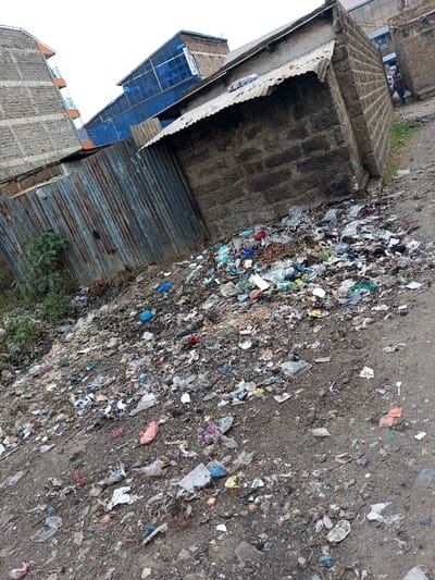 The image displays an unpaved, extensively littered area within Tala ward, Kenya. A prominent accumulation of mixed refuse, consisting primarily of plastic bags, plastic bottles, paper, and assorted domestic waste, covers the dirt ground across the foreground and midground. To the right, a single-story structure constructed from dark grey cinder blocks is present, topped with a sloped, rusted corrugated metal roof. Immediately to its left, a tall, vertical corrugated metal fence, exhibiting significant rust and discoloration, separates the foreground from sparse green vegetation. In the background, an overcast sky is visible above several urban structures: a multi-story building of light-colored brick with orange balcony accents on the far left, and a central building featuring dark blue paneling. A dirt path extends into the distance on the right, where a single individual, dressed in dark clothing, is observed walking away from the camera. No discernible interactions or specific actions are occurring in the immediate foreground.