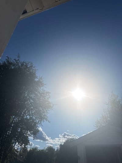 The image shows a bright sun in a clear blue sky, with some scattered clouds. On the left side of the image, there is a portion of a white overhang and some trees with dense foliage. On the right side, the roof of a white building is visible, possibly a garage, with trees in the background. The sun is the dominant light source, creating a lens flare effect. The scene appears to be an outdoor setting in Moelv, Norway, on a sunny day. There are no people or animals visible in the image.