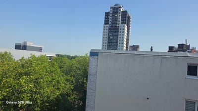 The image shows an urban cityscape with a clear blue sky. In the foreground, a section of a light-colored building with a textured surface dominates the right side, featuring a dark-framed window. To the left of this building, a white brick wall with visible mortar runs vertically.
Behind these structures, lush green trees with dense foliage are a prominent feature. In the middle ground, a tall, modern residential skyscraper with a distinctive facade of white and dark panels, interspersed with balconies, stands out against the sky. Further in the distance, other buildings, including a longer, lower building with a corrugated roof and large purple signage, are visible. The sky is a bright, clear blue, suggesting it is daytime, likely late morning or afternoon, with fair weather.
The scene appears to be a view from a window or elevated position looking out over a city with a mix of residential and commercial architecture, framed by abundant greenery. The overall impression is one of a sunny day in a developed urban area.
The visible text "Galaxy S26 Ultra" is present in the bottom left corner of the image, indicating the device used to capture the photograph.