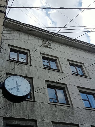 The image is a low-angle shot of a building facade in Saint Petersburg, Russia, with a clock mounted on its side. The building is constructed of light gray, textured stone blocks and features several rectangular windows with dark wooden frames. Overhead, a tangle of black electrical wires crisscrosses against a cloudy sky. A large, round clock face, also with Roman numerals and black hands, is prominently displayed to the left of the frame, its surface reflecting the sky and surrounding architecture. The clock appears to be indicating a time around 11:05. The building's architecture suggests it's an older structure, possibly dating from the Soviet era, given the inscription "ХРОНОТРОН" (Chronotron) visible on the clock face, which was a Soviet brand of clocks. The overall impression is one of an urban environment with a vintage feel.