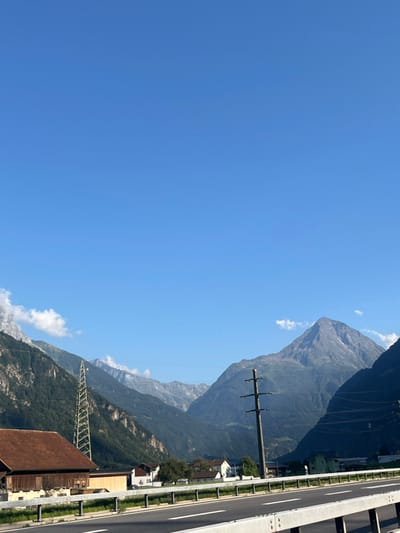 The image displays a panoramic daytime view of a mountainous landscape in the vicinity of Erstfeld, Switzerland, as seen from an elevated position on a highway. The sky is uniformly clear blue with minimal, scattered white clouds primarily near the mountain peaks. In the immediate foreground, a grey metal crash barrier, typical of highway infrastructure, runs along the right edge, bordering an asphalt multi-lane road featuring white lane markings.
To the left and below the elevated highway, a traditional two-story wooden building with a reddish-brown gabled roof is partially visible. Further back on the left, a tall, lattice-structured metal power transmission tower stands against a densely forested mountain slope. In the midground, a cluster of residential buildings with white facades and dark roofs is discernible among green foliage, indicating a small settlement. A dark utility pole with multiple horizontal cross-arms is prominently positioned to the right of these structures, supporting overhead power lines that extend horizontally across the scene.
The background is dominated by successive layers of mountains. The closest range is heavily covered in dark green forest, while the more distant peaks, including a distinct triangular-shaped summit on the right, appear rockier with less vegetation. No individuals, vehicles, or animals are present within the depicted scene. The environment is uniformly illuminated by direct sunlight, indicating a clear weather condition.